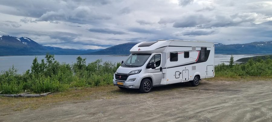 De camper op een grindvlakte voor een meer en bergen met sneeuwresten